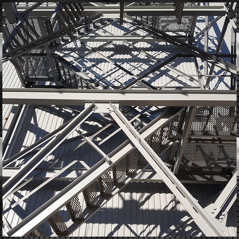 Industrial grey metal fire escape with beams and mesh steps casting geometric shadows on a sunlit, corrugated metal wall.