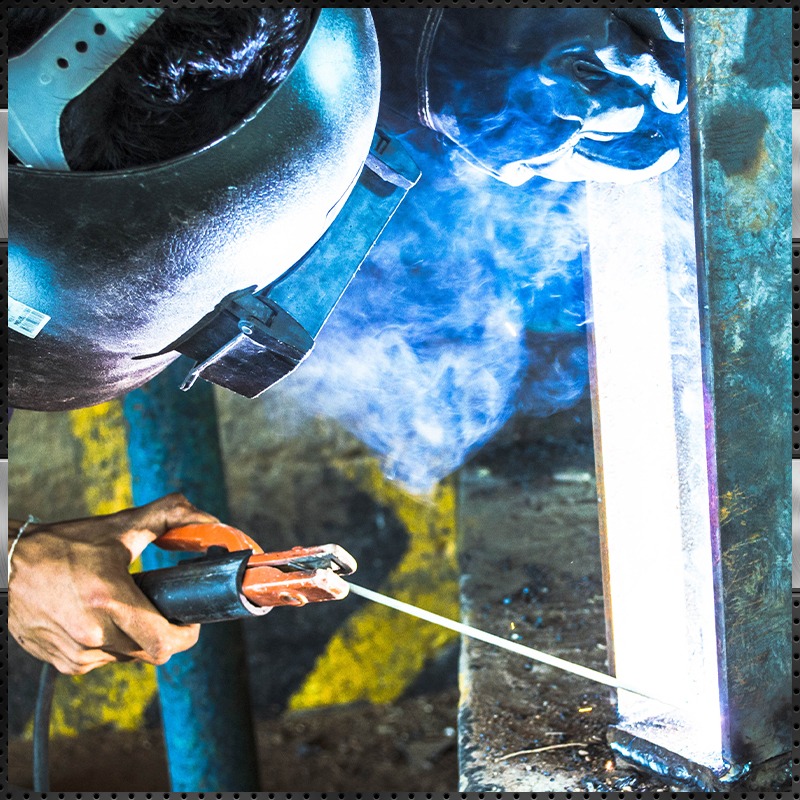 Industrial welder performing stick arc welding on a steel I-beam, with bright sparks, glowing arc light, and blue smoke capturing intense metal fabrication and structural steel construction work.