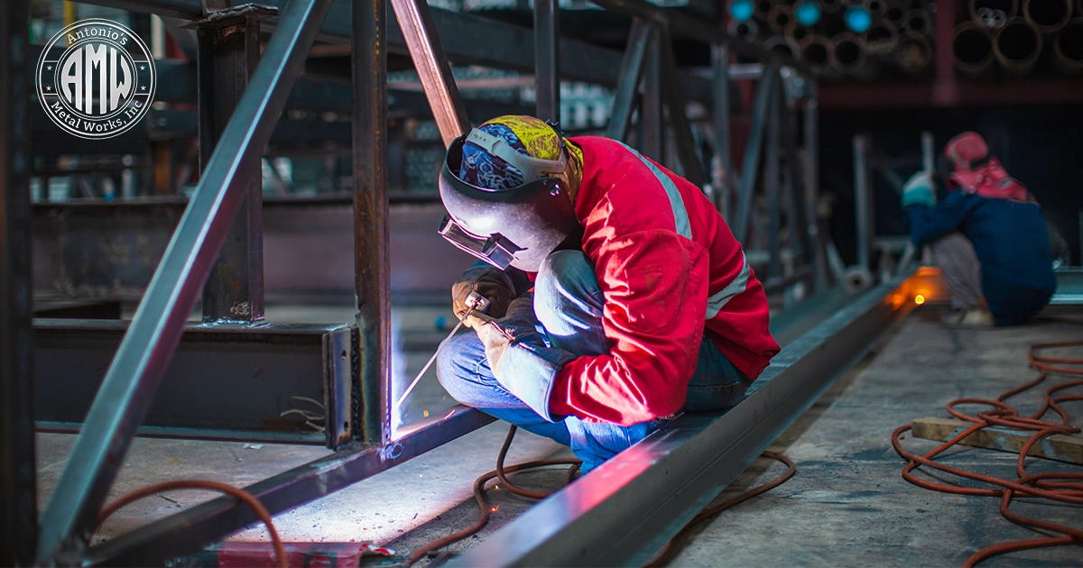 A low angle photo of a welder in a red jacket welding steel beams in a dark fabrication shop.
